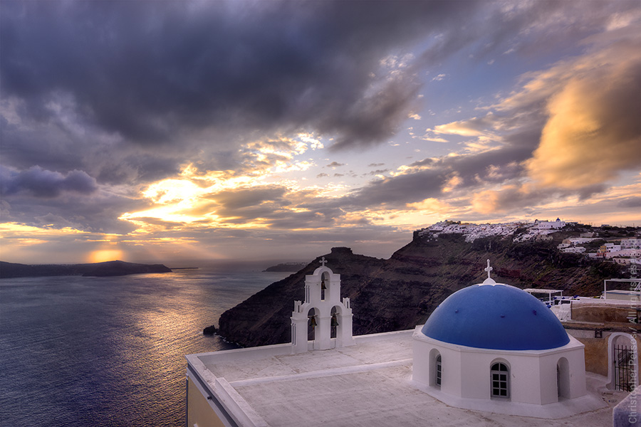 Sunset behind clouds with a blue dome church high above the water on Santorini by Christopher Petsos