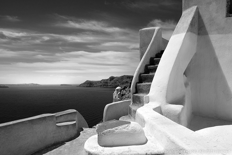 Black and white photography of whitewashed stairs above the water with bright sunshine and a dark sea by Chris Petsos