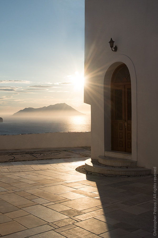 Fine art photography print by Christopher Petsos: The sunset on Milos, Greece seen from a terrace above the sea with island architecture