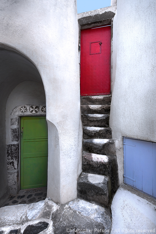 Fine art print by Chris Petsos of three colorful doors and whitewashed walls with steps in Santorini, Greece