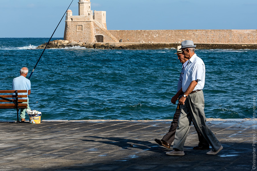 Two Greek men in hats walk along the water in Chania with the lighthouse and a fisherman holding a pole